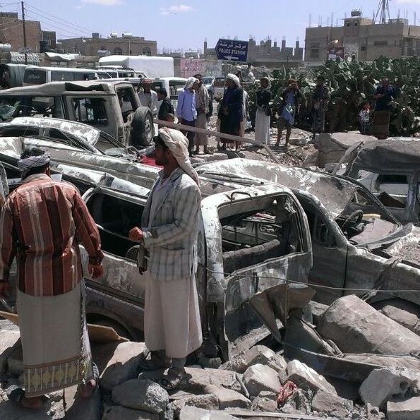 The aftermath of a bombing by the Saudi-led coalition in Yemen © Almigad Mojalli/IRIN Der Einschlagsort einer Bombe ist zu sehen. Komplett ausgebrannte Autos, verkohlte Rest und schwarze Erde, Trümmer. Dazwischen stehen einige Männer.