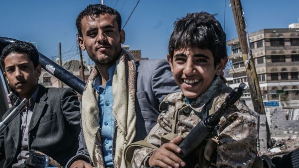 Child soldiers are common in Yemen, with the Houthis among several groups to use them ©Mohammed Al-Ariqi/IRIN Ein Junge sitzt neben einem Erwachsenen auf einer Ladefläche eines Pick-Up und hat ein Sturmgewehr in der Hand. Er lächelt. Der Mann schaut ihn interessiert an. Im Hintergrund sind Ruinen zu sehen. Ganz links sitzt ein weiterer etwas älterer junger Man