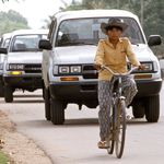 Eine Fahrradfahrerin in Kambodscha fürht einen UNTAC-Autokonvoy an. Eine Fahrradfahrerin fährt vor einer langen Reihe an weißen Autos.